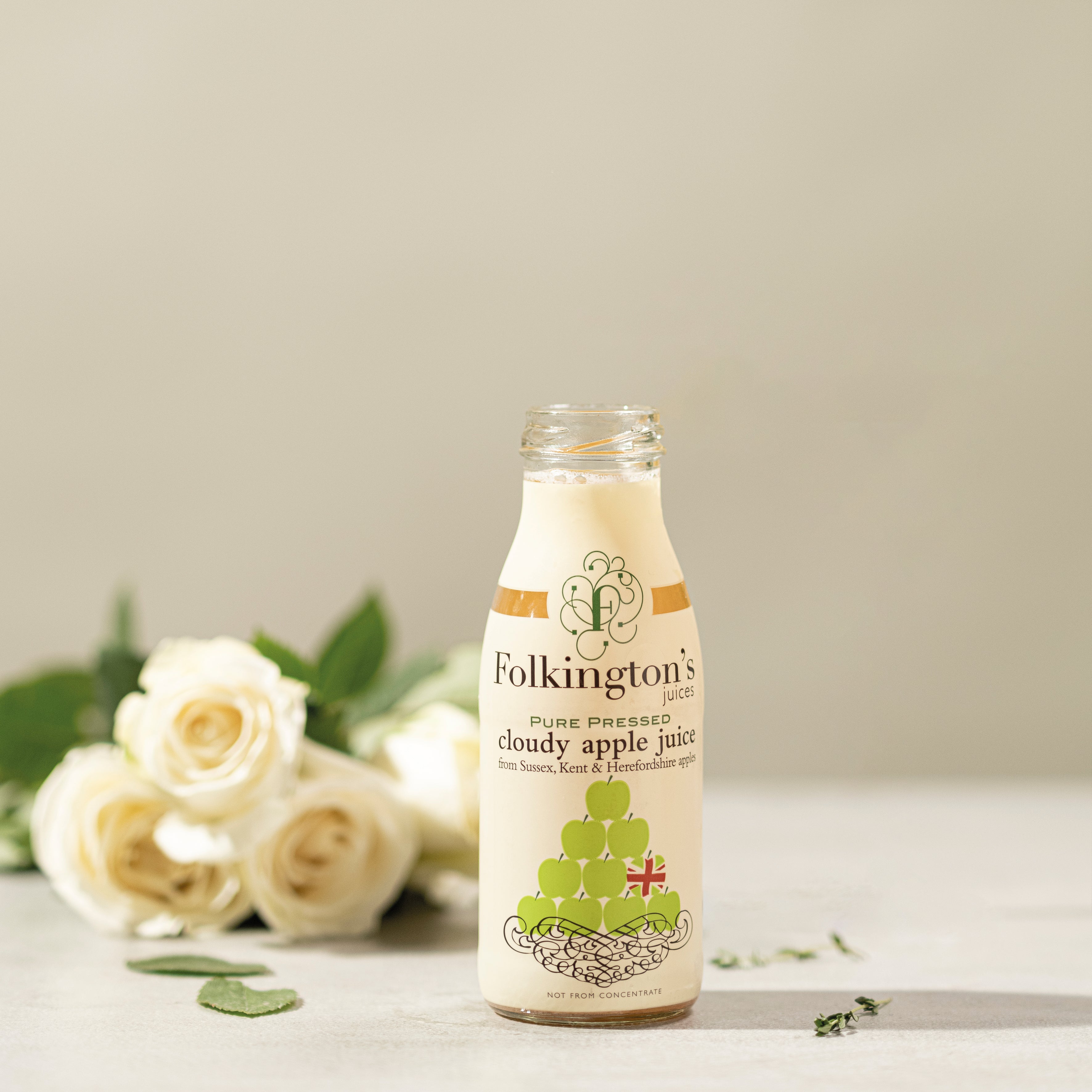 Folkington's Cloudy Apple Juice in a clear glass bottle with a white and gold label, beside white roses.