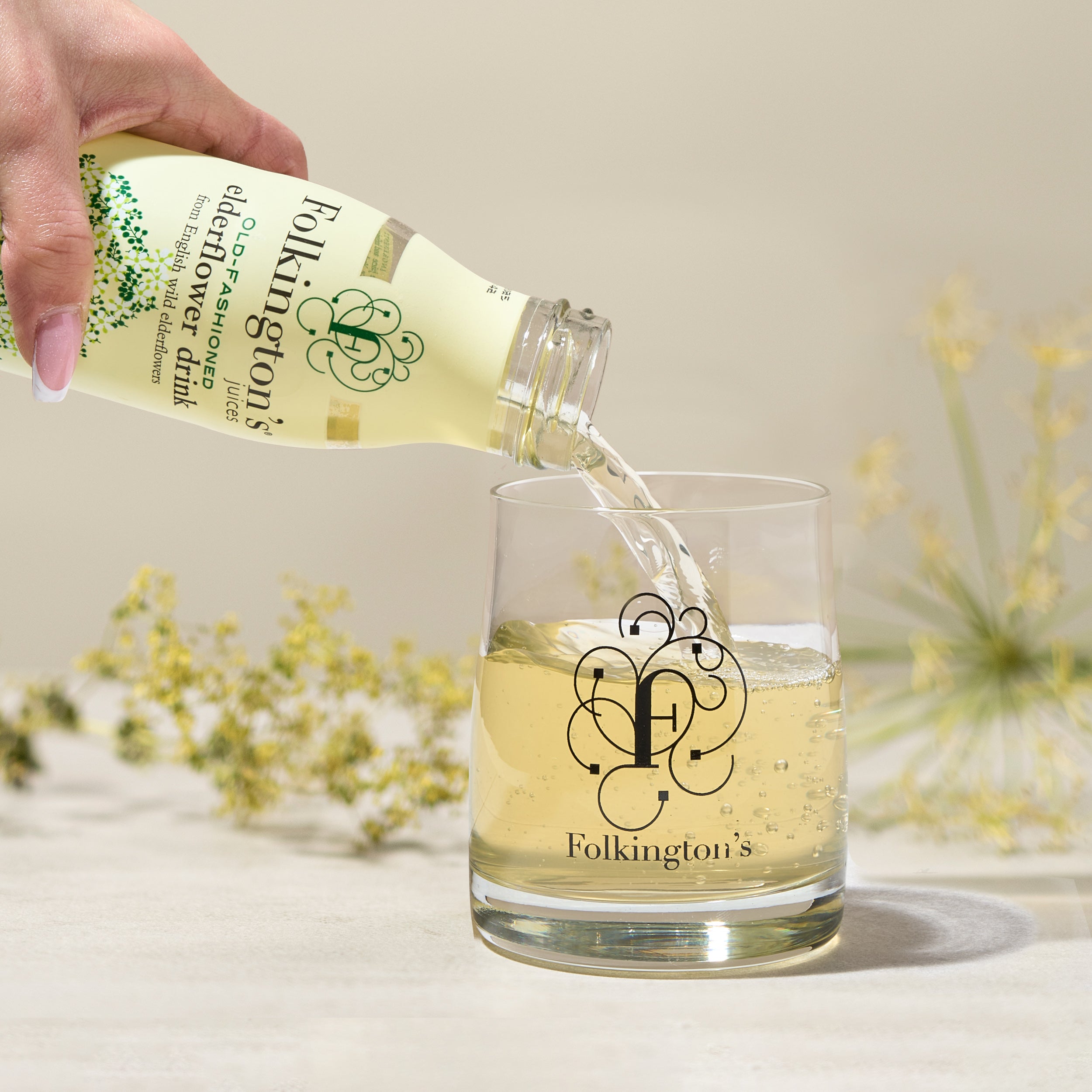 Hand pouring Folkington's elderflower drink from a 250ml bottle into a clear glass with the brand logo.