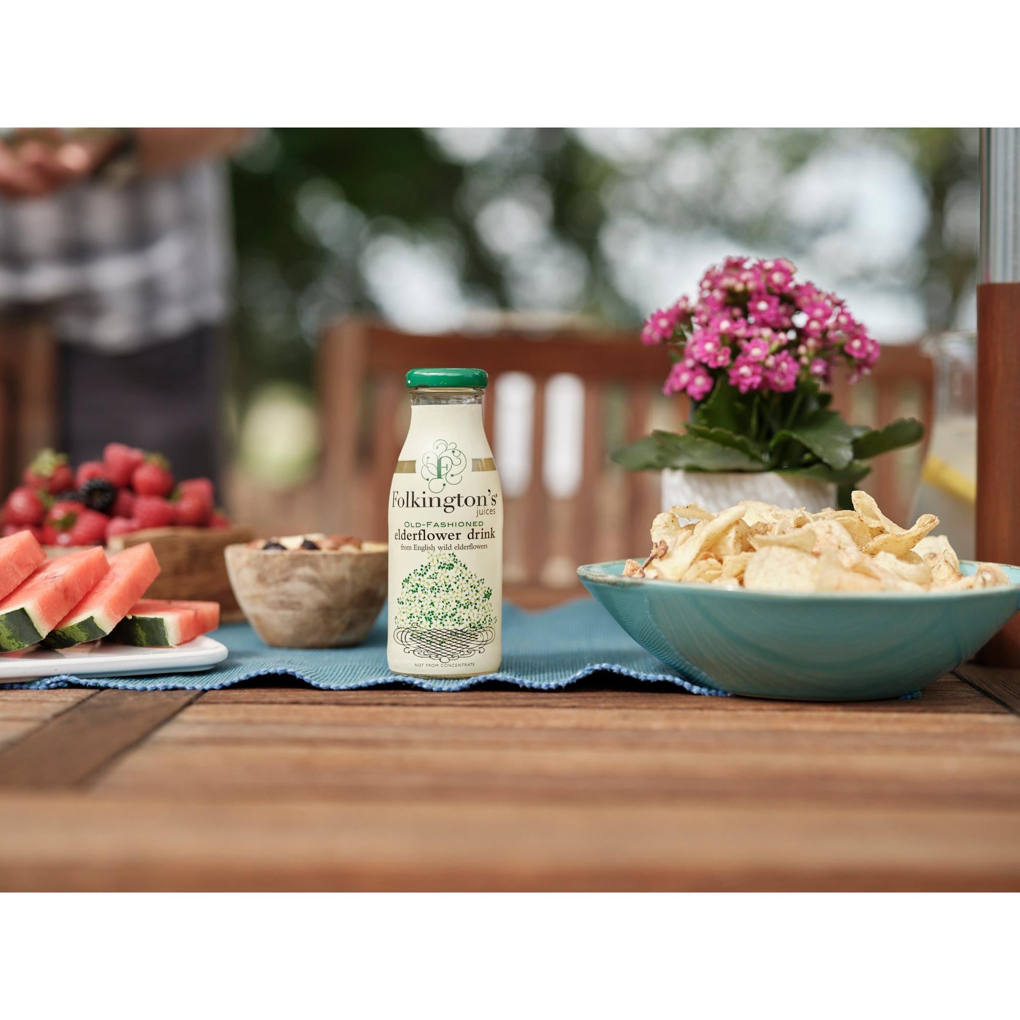 Folkington's Old-Fashioned Elderflower Drink in a clear glass bottle with green cap, on a wooden table with snacks.