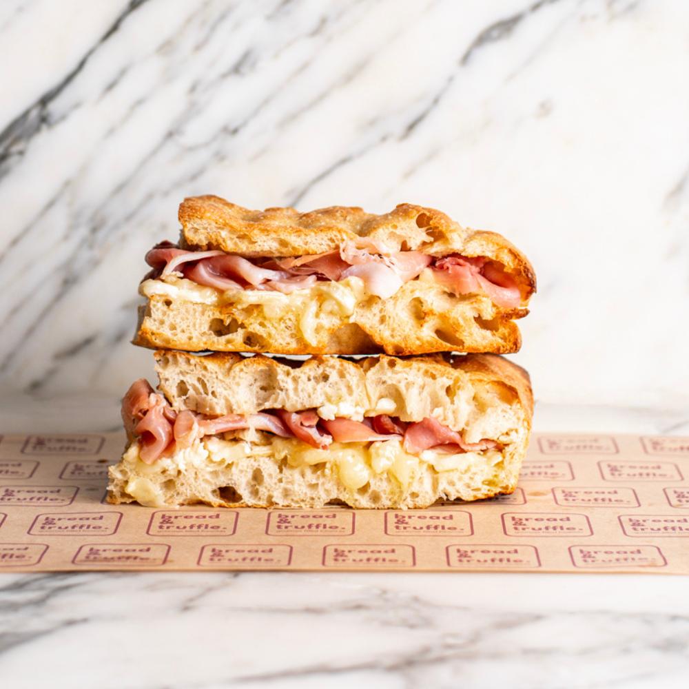 Stacked focaccia sandwich with speck, creamy brie, and golden crust on branded paper.