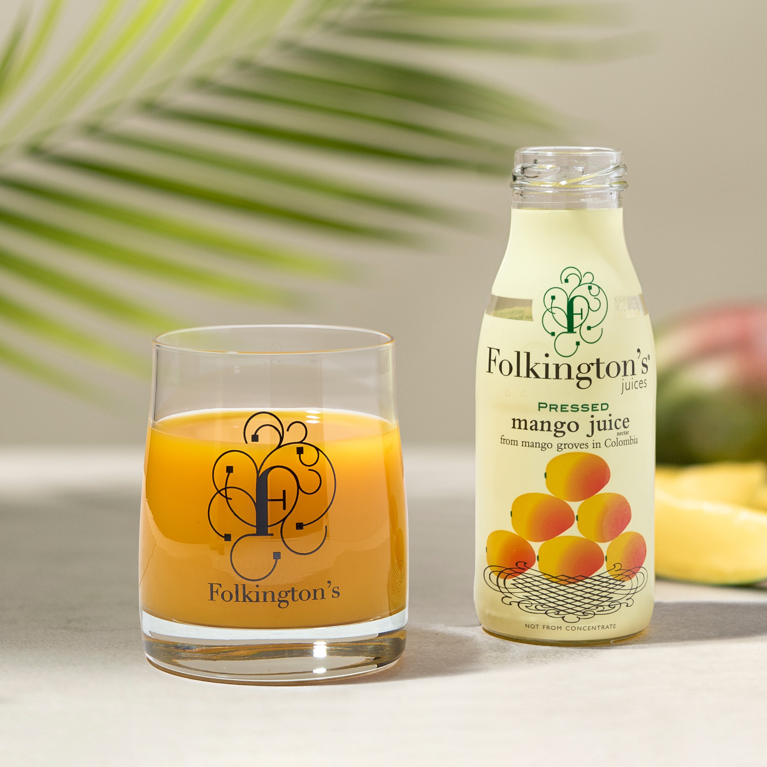 Folkington's mango juice bottle and a glass of bright orange mango nectar, with a palm leaf background.