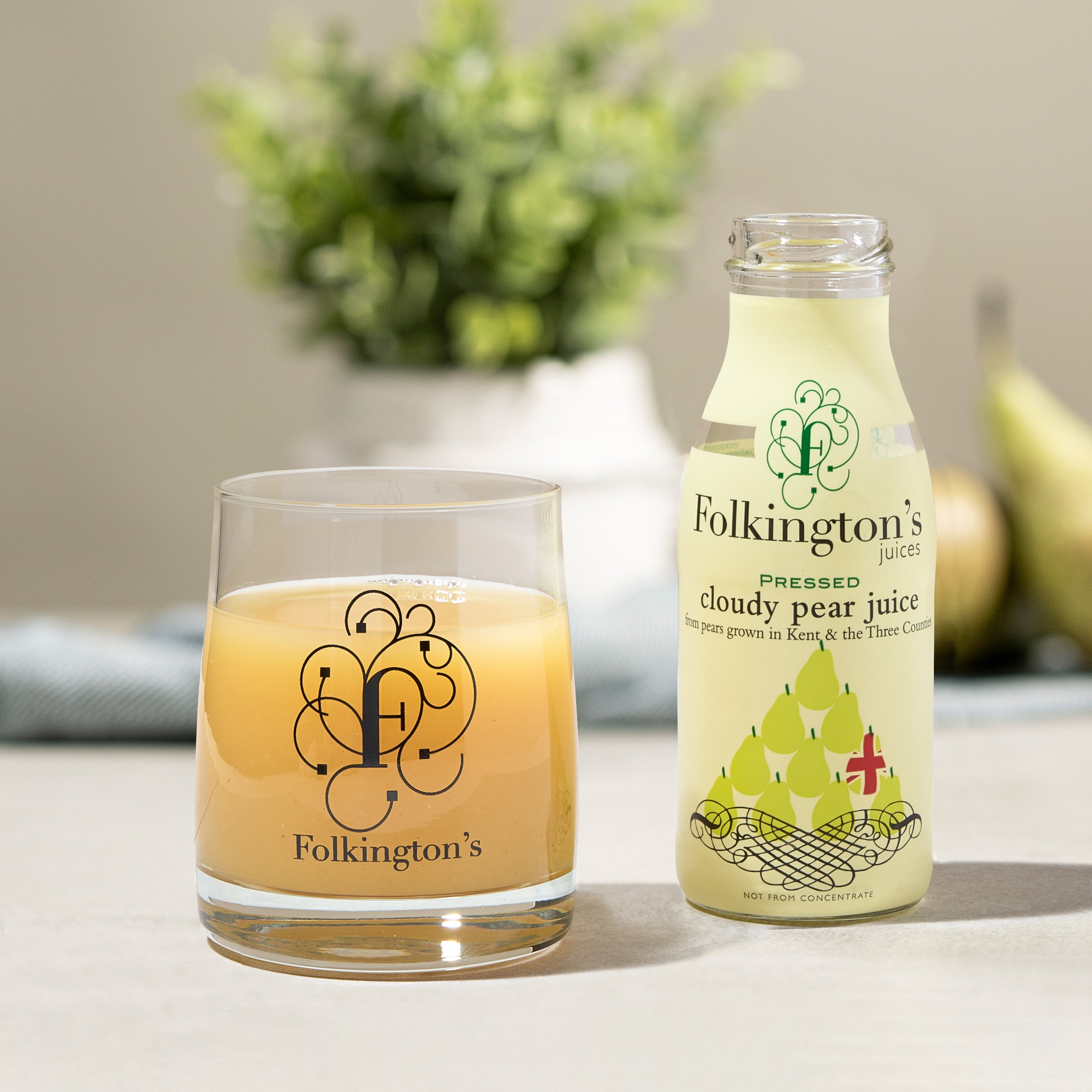 Folkington's Cloudy Pear Juice bottle and a glass filled with the golden juice, with pears in the background.