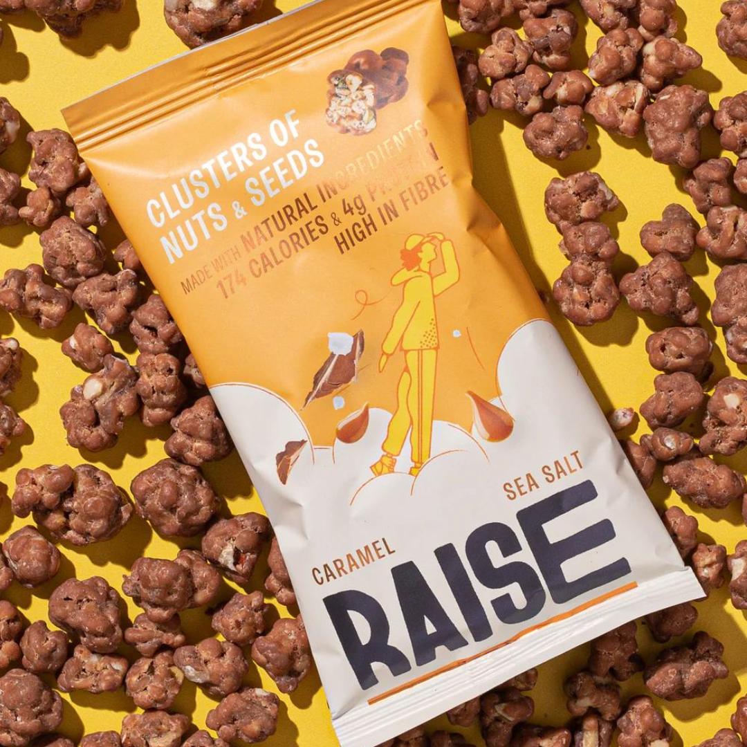 Raise Snacks Salted Caramel pouch on a yellow background, surrounded by chocolate-coated nut and seed clusters.