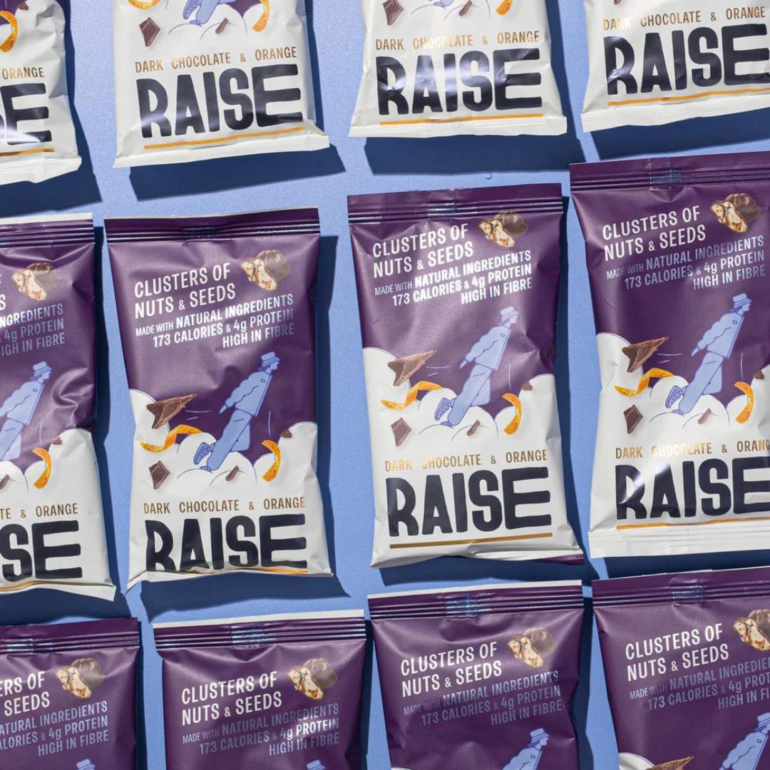 Multiple purple and white Raise Snacks Dark Chocolate & Orange Zest pouches with nuts, seeds, and chocolate graphics.