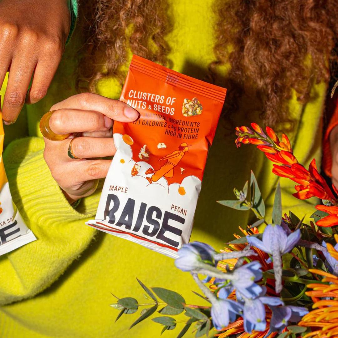 Hand holding an orange Raise Snacks Maple Pecan pouch with nuts, seeds, and a person running design.