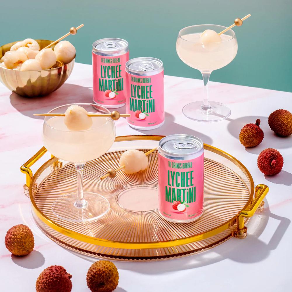 Three pink Lychee Martini cans with two cocktails garnished with lychee on a gold tray.