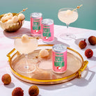 Three pink Lychee Martini cans with two cocktails garnished with lychee on a gold tray.