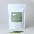 White Marna ceremonial grade matcha pouch with green label, next to a sprinkle of vibrant green matcha powder.