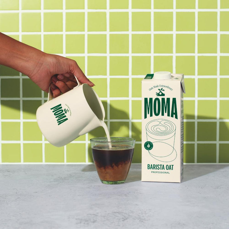 Hand pouring MOMA Barista Oat milk from a cream pitcher into a coffee glass, next to a 1L carton.