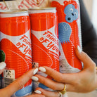 Hands holding three vibrant red and white Fountain of Youth coconut water cans with a blue monkey logo.