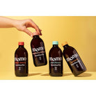 Hand holding a MOMO raspberry-hibiscus kombucha bottle, with ginger-lemon, elderflower, and turmeric bottles on a yellow background.