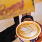 Hand holding a Bonsoy latte with intricate leaf art, a yellow Bonsoy carton in the background.