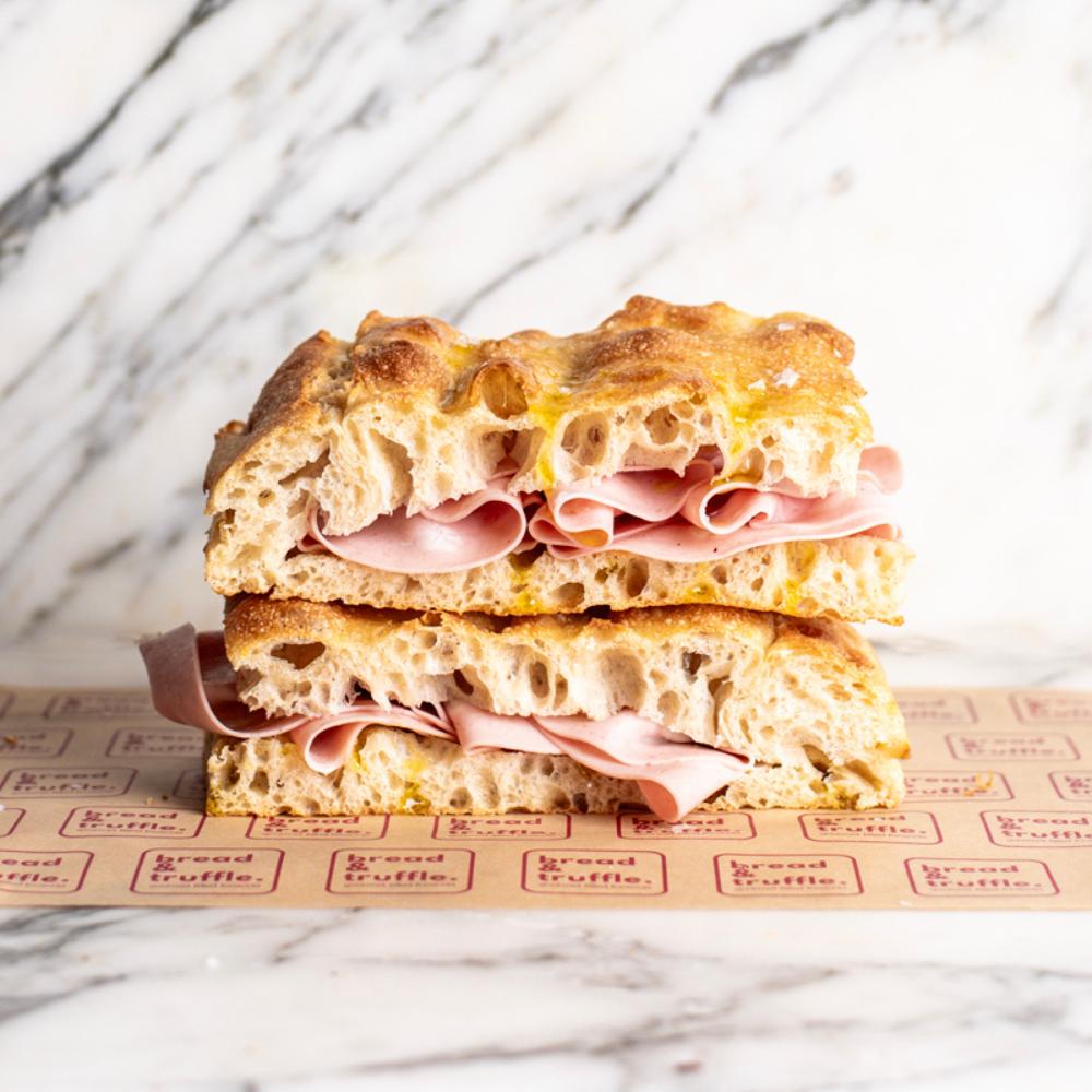 Stacked focaccia sandwich with pink Italian ham and golden crust, on branded brown paper.