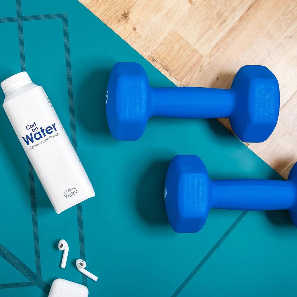 White Carton Water carton with sugarcane cap, next to blue dumbbells and white earbuds on a teal yoga mat.