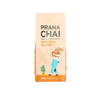 Prana Chai Original Blend 1kg bag, beige with orange base, featuring a person holding a chai cup and a plant.