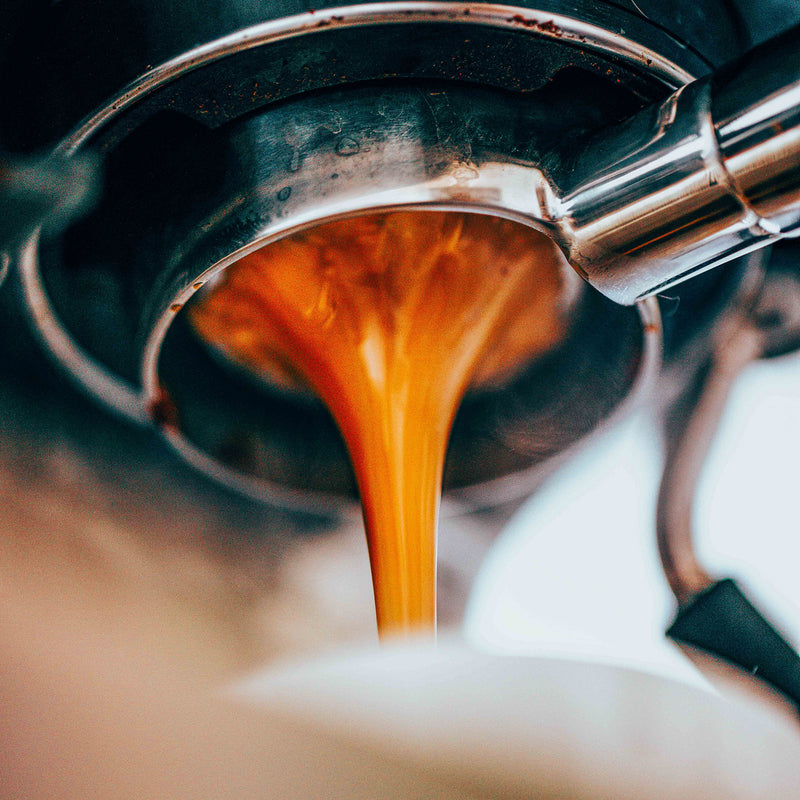 Close-up of rich, dark orange espresso pouring from a shiny chrome portafilter into a white cup.