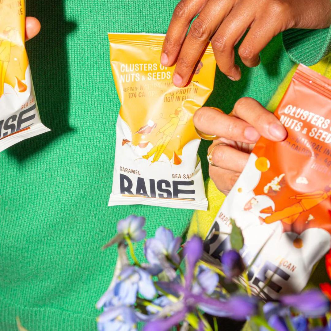 Hands holding yellow and orange Raise Snacks Salted Caramel clusters pouches against a green sweater.