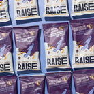 Multiple purple and white Raise Snacks Dark Chocolate & Orange Zest pouches with nuts, seeds, and chocolate graphics.