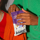 Hands opening a purple and white Raise Snacks pouch, Dark Chocolate & Orange flavor, with abstract design.