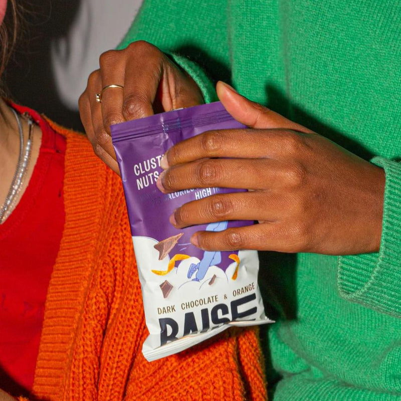Hands opening a purple and white Raise Snacks pouch, Dark Chocolate & Orange flavor, with abstract design.