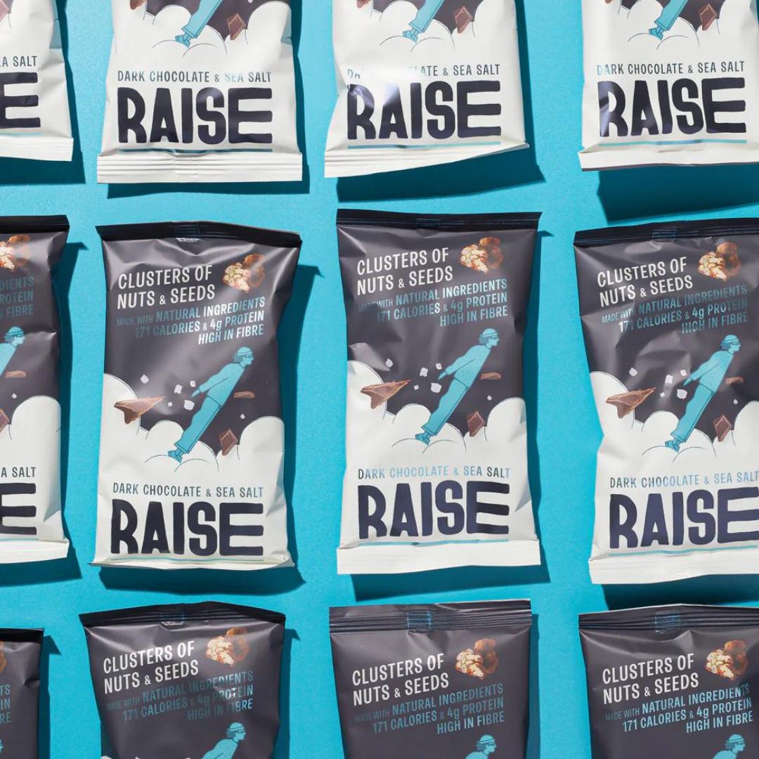 Multiple Raise Snacks Dark Chocolate & Sea Salt pouches, dark gray with white branding, arranged on a blue surface.
