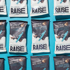 Multiple Raise Snacks Dark Chocolate & Sea Salt pouches, dark gray with white branding, arranged on a blue surface.